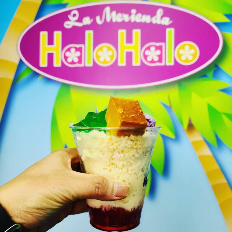 Me holding a cup of halo-halo in front of a wall with the store name: La Merienda HaloHalo.